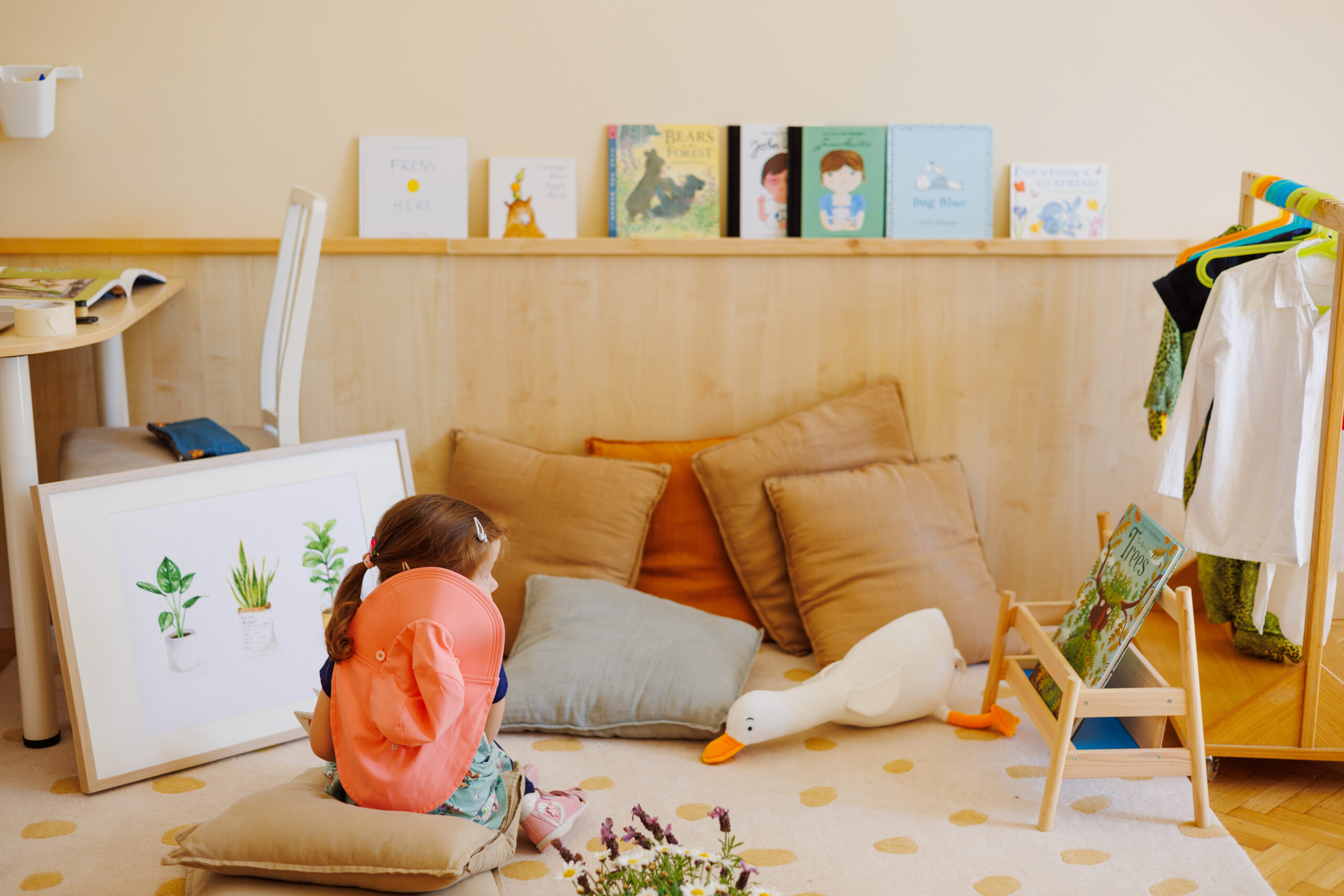 Kindergarten reading corner with a child, pillows, books at eye level, and neatly hung dress-up costumes